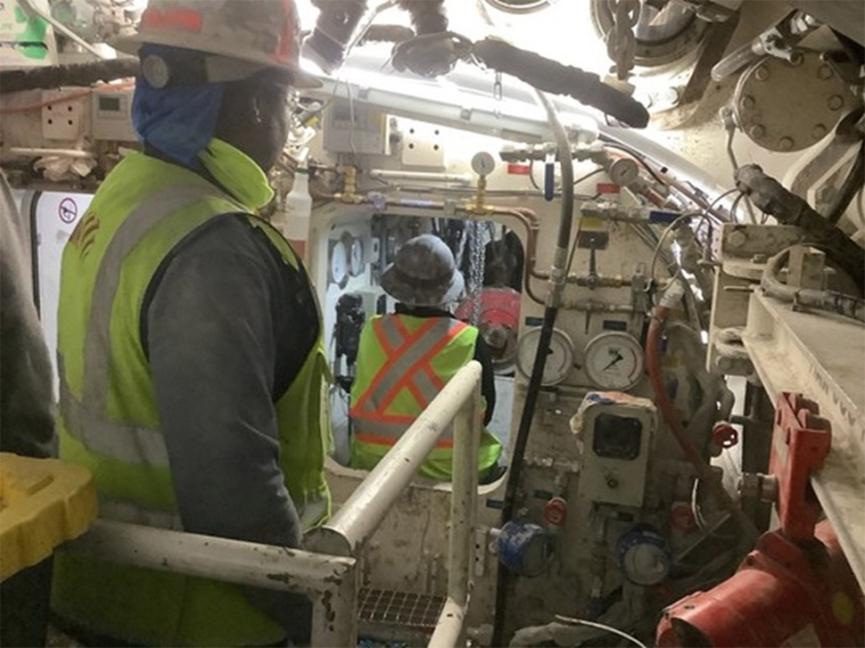 Two people in bright yellow vests and hard hats are inside the tunnel boring machine and surrounded by various gages and electricity conduits. One of them is sending a new cutterhead tool through a small door that leads to the front of the machine. 