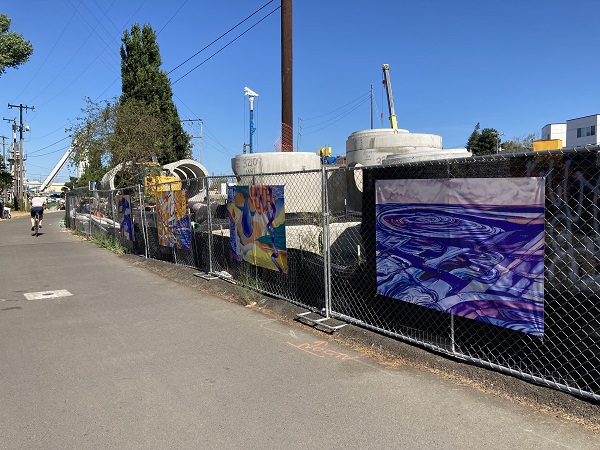 Four vinyl banners that showcase water-related digital paintings. They are secured to a chain link fence with a black construction screen