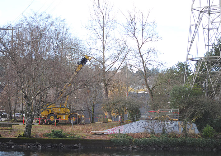 A yellow excavator surrounded by orange cones is situated next to trees at the future site of Queen Anne vertical shaft at the end of W Ewing St from across the Ship Canal in Fremont.