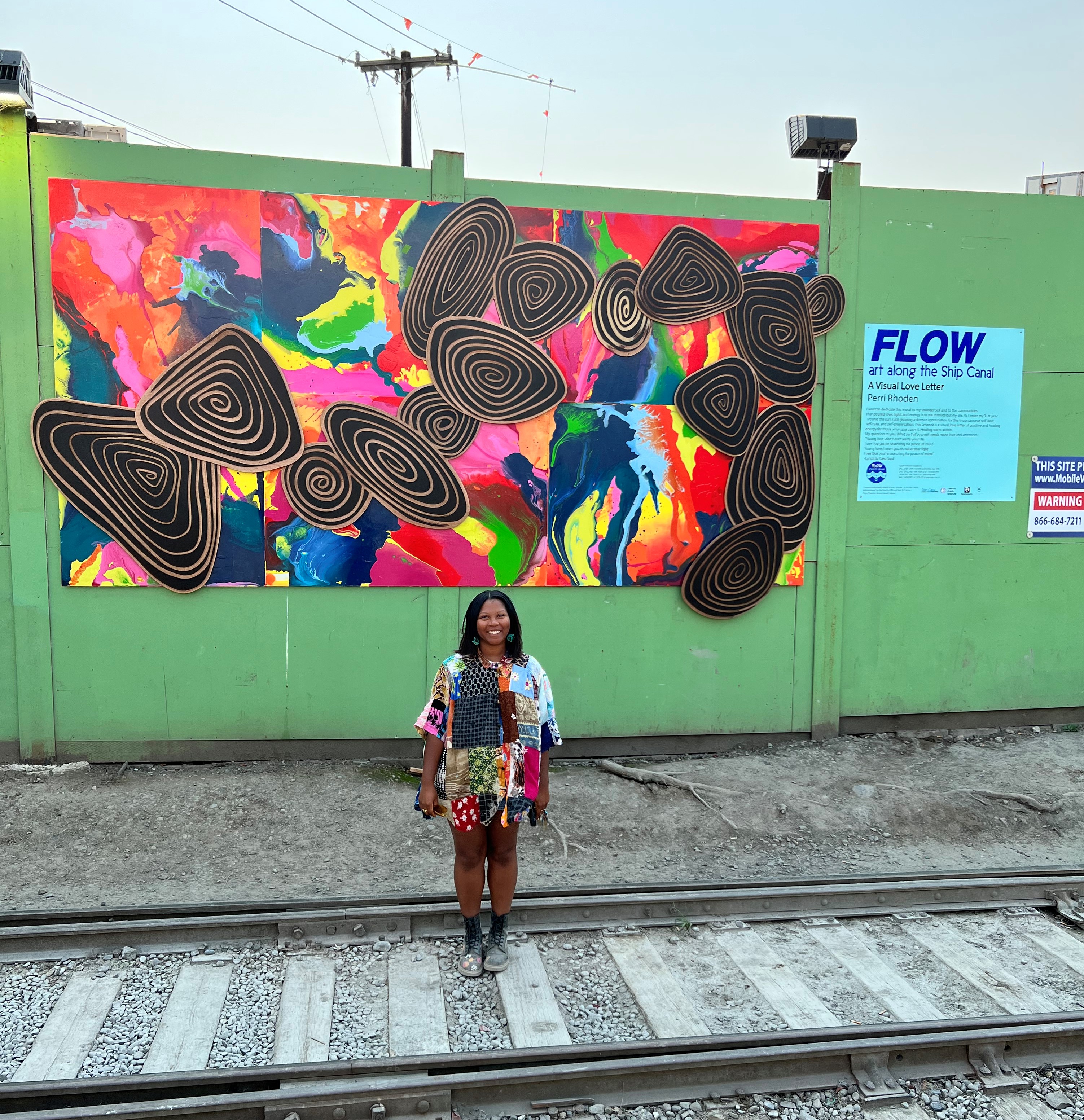 Artist, Perri Rhoden, stands in front of their mural. The Mural is brightly multicolored with black and gold spirals throughout the piece. 