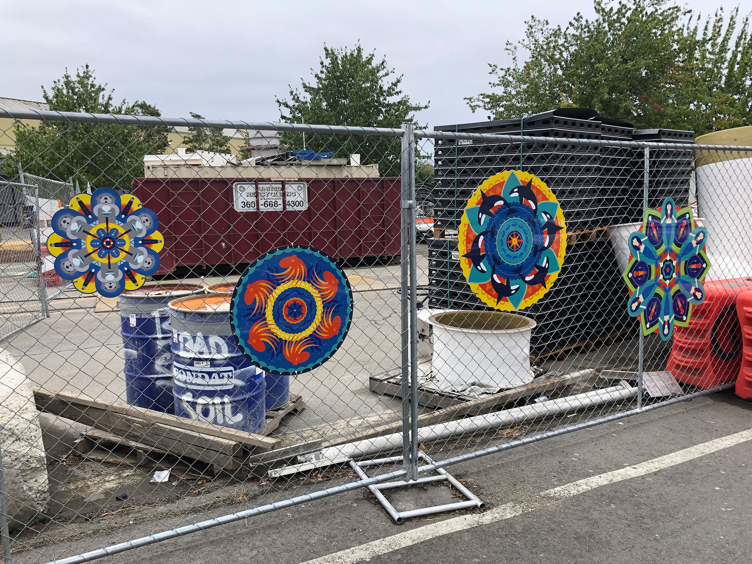 Four kaleidoscope-style medallions that highlight aquatic icons using primarily blue, red and yellow colors. They are secured to a chain link fence with construction materials in the background.