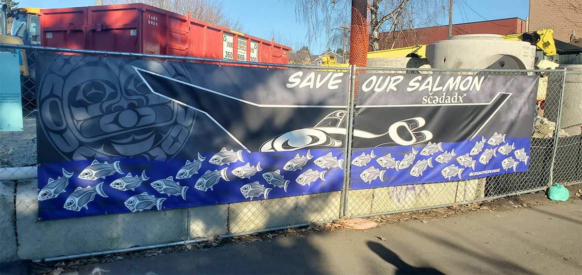 This photo shows a banner with native-style salmon swimming down a river toward the moon. The river also has a canoe floating on top of it with “save our salmon” written above the canoe. 