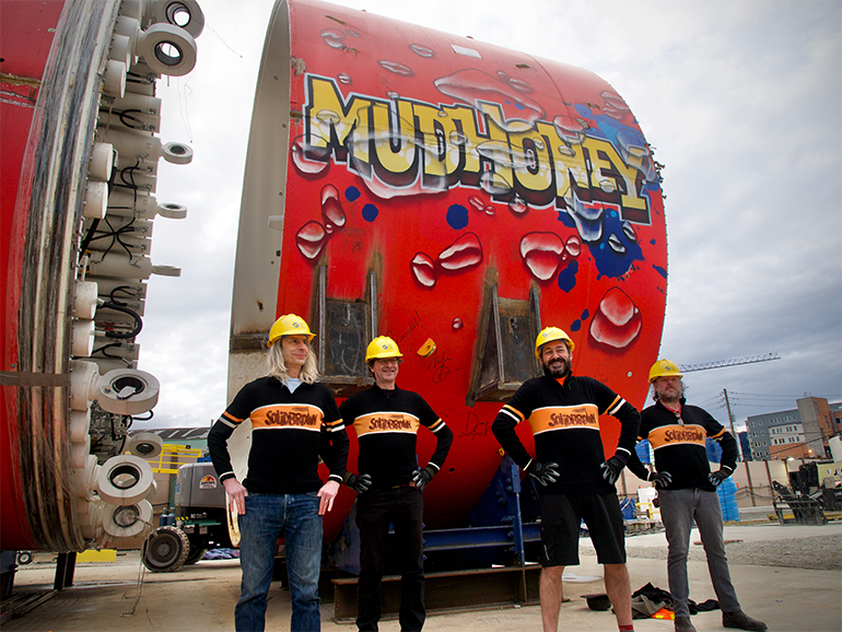 Four individuals wearing sweaters and hardhats stand in front of a tunnel boring machine that has the word MudHoney drawn in yellow and black letters